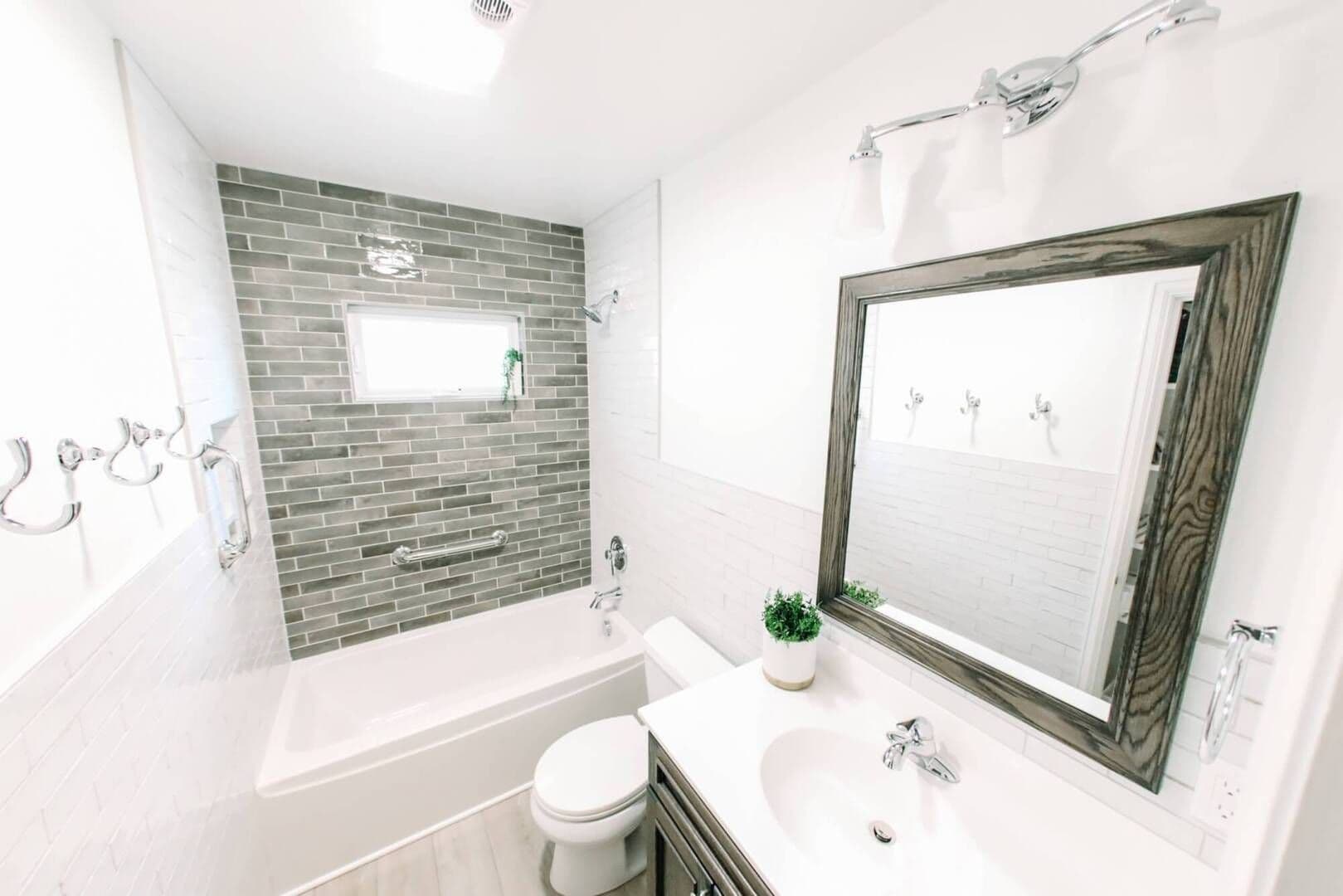 aerial view of small white bathroom with gray tiling on opposite wall above white bathtub and gray cabinetry. White furnishings and silver accents, counter top and toilet on the right with gray wood framed mirror above sink