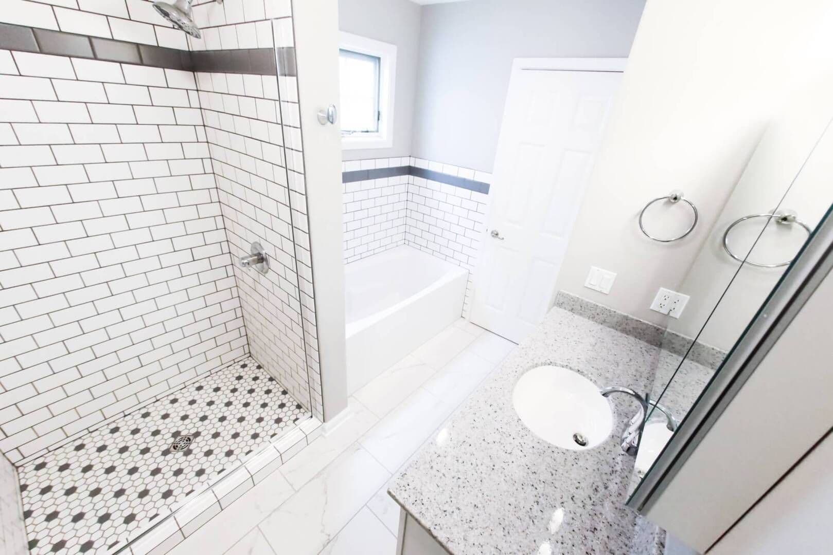 aerial view looking down from toilet. Sink/countertop to the right with black/white tiled shower across from toilet and white bathtub next to it. Silver fixtures/accents throughout